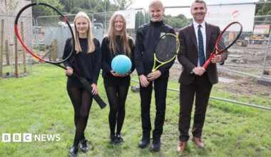 A group of four people, three of them younger and all dressed in black and one older in a suit and tie. They are carrying tennis racquets - one of them very large - and a large blue tennis ball.