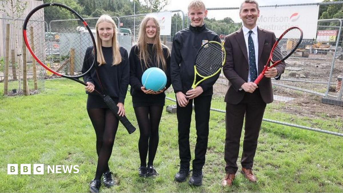 A group of four people, three of them younger and all dressed in black and one older in a suit and tie. They are carrying tennis racquets - one of them very large - and a large blue tennis ball.