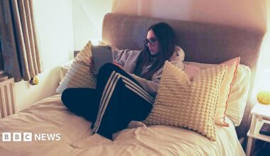 Anna-May, who has long brown hair and wears glasses, is propped up on lots of cushions on her bed, which has a light grey headboard. She is watching an iPad which she is holding in her hands, wearing tracksuit bottoms and a hoodie. There are bedside lights on tables on either side of her bed.