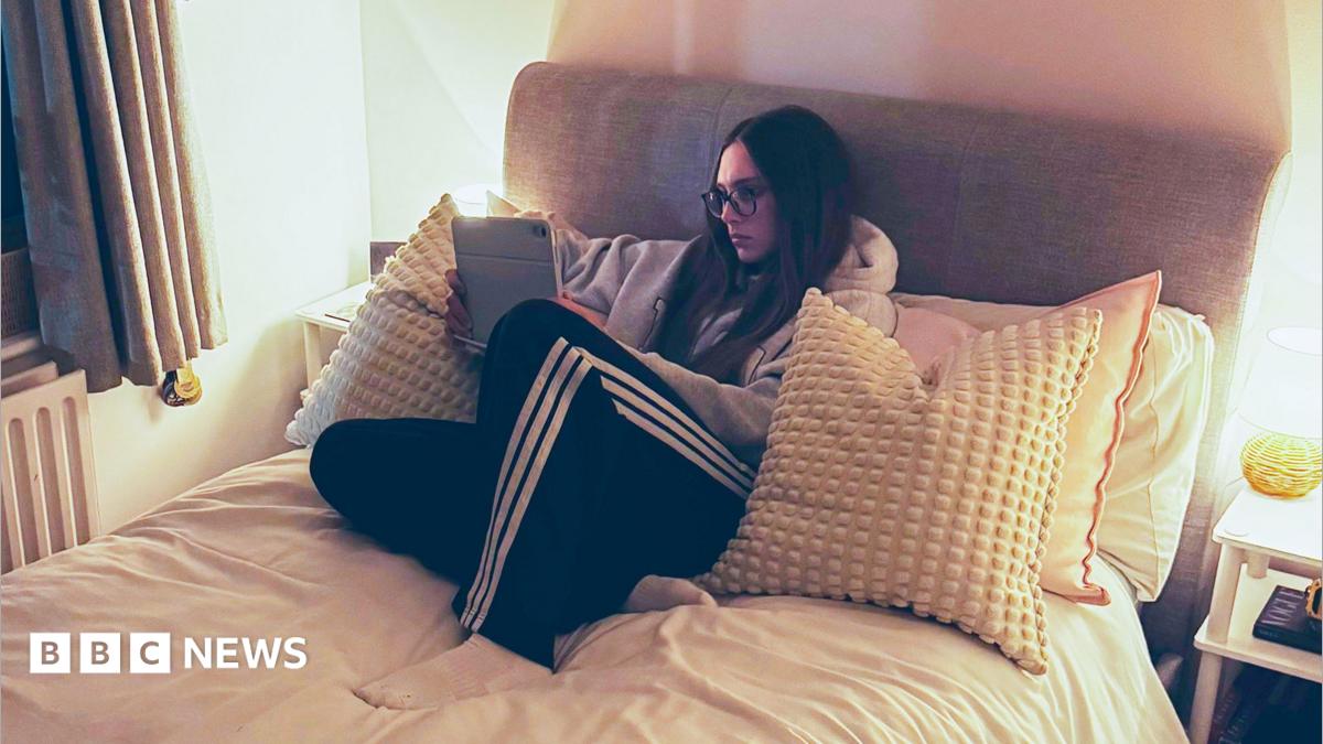 Anna-May, who has long brown hair and wears glasses, is propped up on lots of cushions on her bed, which has a light grey headboard. She is watching an iPad which she is holding in her hands, wearing tracksuit bottoms and a hoodie. There are bedside lights on tables on either side of her bed.