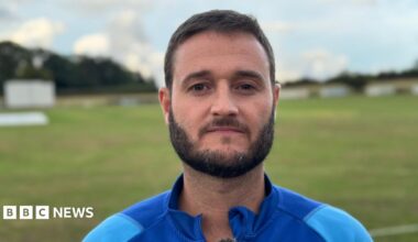 A man with black hair and beard, wearing a light blue tracksuit with a blue collar