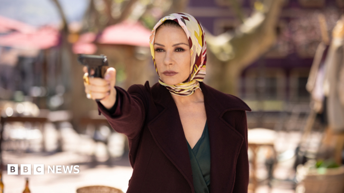Catherine Zeta-Jones holding a gun up, wearing a scarf around her head and a maroon jacket in the Kill Jackie series.