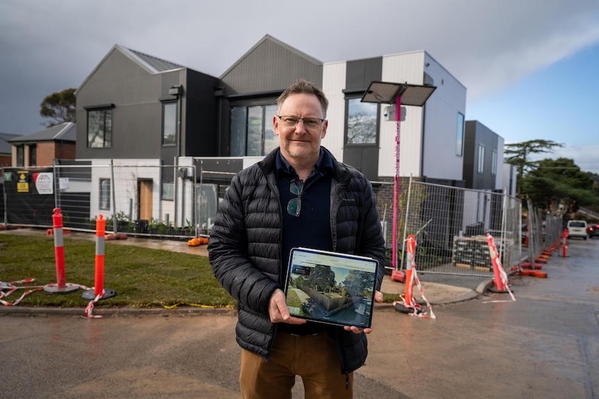 A man in a puffer jacket holds an iPad outside a suburban construction site