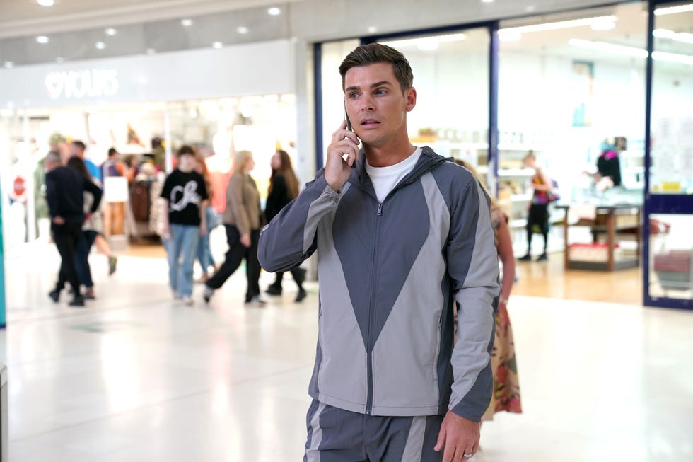 hollyoaks ste hay man in tracksuit talking on a phone in a busy mall.