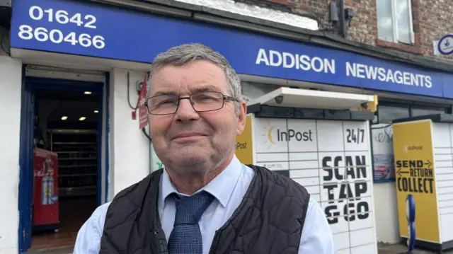 A man stands outside a shop. He is wearing a pale blue shirt and tie. He has glasses and short, cropped grey hair.