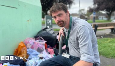 A wide shot of a man crouching down next to a green charity donation bin, with bags full of clothes and rubbish on the floor beside the bin. He has brown hair, a beard and is wearing a grey shirt. A patch of green grass, a wooden bench and a roadway can be seen in the background, which is out of focus.