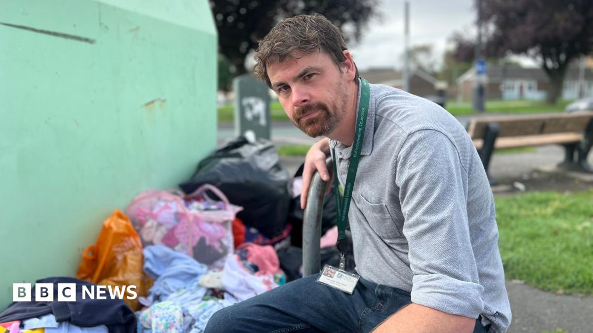 A wide shot of a man crouching down next to a green charity donation bin, with bags full of clothes and rubbish on the floor beside the bin. He has brown hair, a beard and is wearing a grey shirt. A patch of green grass, a wooden bench and a roadway can be seen in the background, which is out of focus.