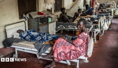 A woman sits at the foot of a bed that has a male patient on it. There are filled beds behind her.