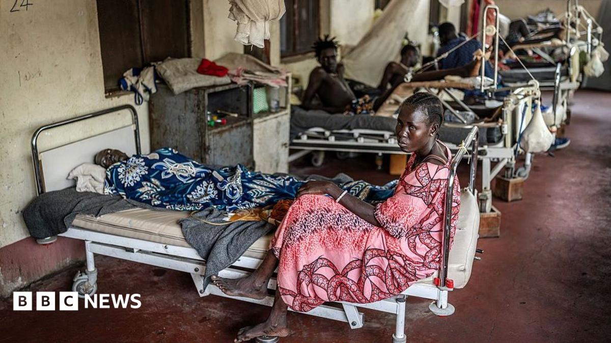 A woman sits at the foot of a bed that has a male patient on it. There are filled beds behind her.