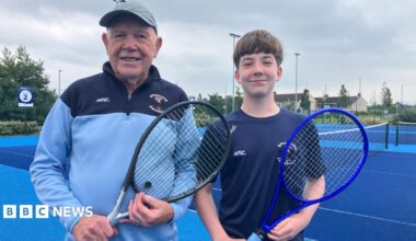 A man wearing a grey hat and blue and navy 1/4 zip and teen boy with brown hair wearing a navy top. They are both holding tennis rackets