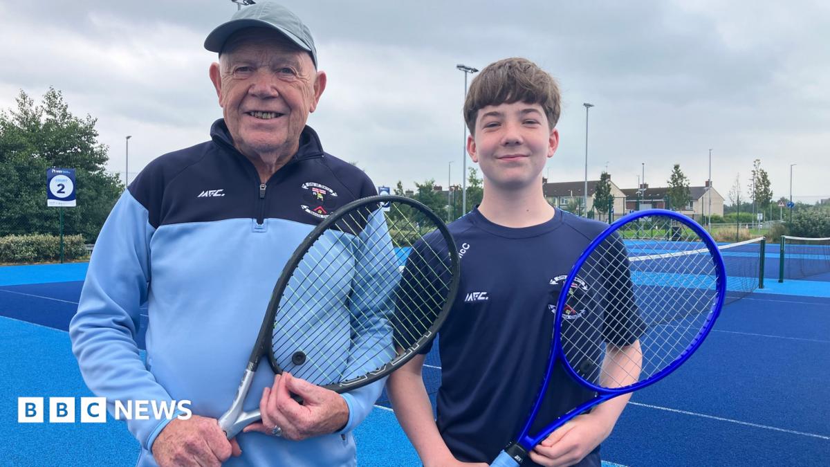 A man wearing a grey hat and blue and navy 1/4 zip and teen boy with brown hair wearing a navy top. They are both holding tennis rackets