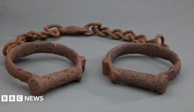 Two iron shackles consisting of iron loops joined together by a length of iron chain. The iron is heavily rusted. These would have been used to restrain enslaved African people forcibly transported to North America and the Caribbean.