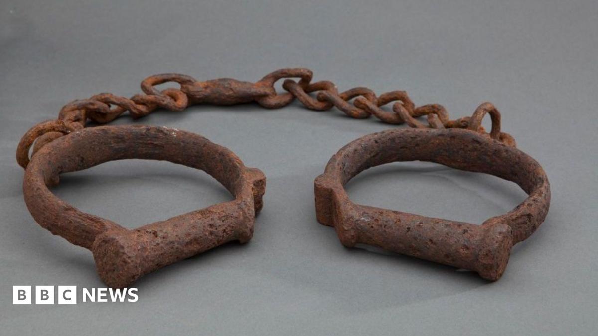 Two iron shackles consisting of iron loops joined together by a length of iron chain. The iron is heavily rusted. These would have been used to restrain enslaved African people forcibly transported to North America and the Caribbean.