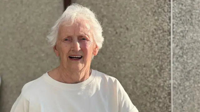 Maureen Gill is smiling as she stands in from of a pebble-dash house. She has short white hair and is wearing a cream-coloured top.