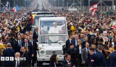 Pope Leo smiles and holds his arm up in his popemobile. Crowds can be seen waving flags and gesturing towards the pope. Some are holding up their phones or cameras to take pictures