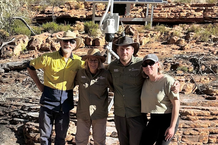 Two men and women, all in outdoor gear and smiling as they stand in front of an outback rock formation.
