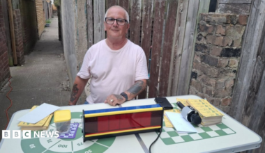 A grey-haired man wearing black glasses. He has tattoos and is wearing a white T-shirt. He is sitting by a table with bingo cards on it, along with a microphone and an electronic display. The table is white with a green check print along with a circle with numbers in it. A narrow alleyway runs behind him.