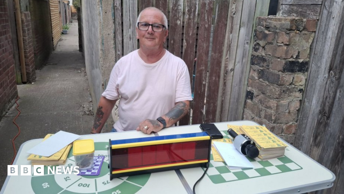 A grey-haired man wearing black glasses. He has tattoos and is wearing a white T-shirt. He is sitting by a table with bingo cards on it, along with a microphone and an electronic display. The table is white with a green check print along with a circle with numbers in it. A narrow alleyway runs behind him.