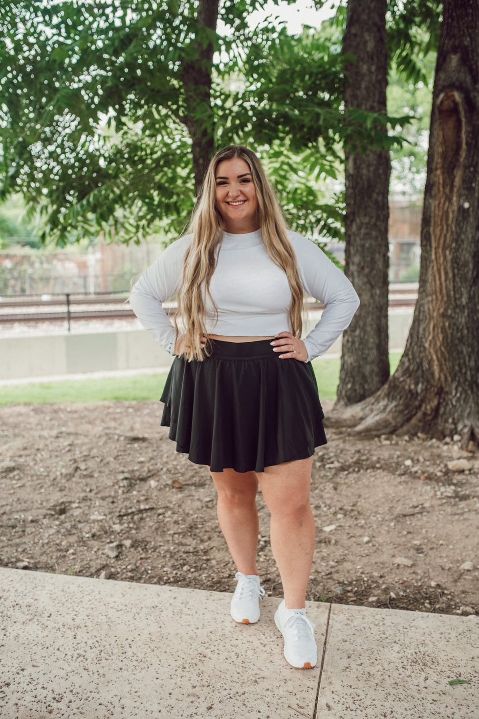 Woman smiling outdoors in a white long-sleeve top and a black skirt, standing among green trees.