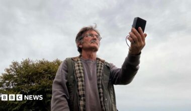 Desmond Consitt wears a grey sweatshirt and green padded gillet. He is holding a rectangular black bat detector in his hand, pointed towards the sky. He has thick grey hair, a moustache and glasses.