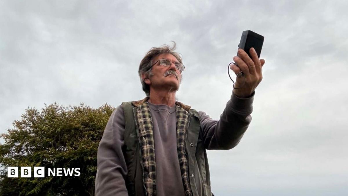Desmond Consitt wears a grey sweatshirt and green padded gillet. He is holding a rectangular black bat detector in his hand, pointed towards the sky. He has thick grey hair, a moustache and glasses.