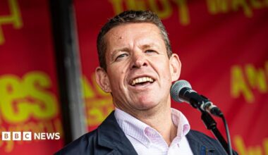 Rhun ap Iorwerth, who has short grey hair and is wearing a salmon pink and white check short and navy blue jacket, speaking into a microphone with red and yello "Yes Cymru" branding behind him.