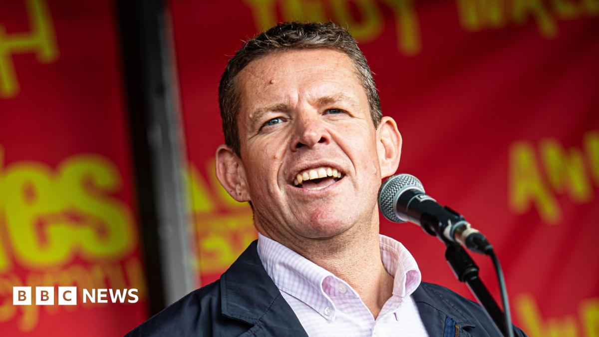 Rhun ap Iorwerth, who has short grey hair and is wearing a salmon pink and white check short and navy blue jacket, speaking into a microphone with red and yello "Yes Cymru" branding behind him.