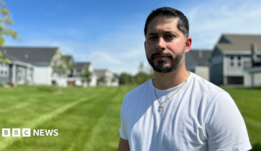 Gerald Bourguet, a white man standing in a field of a manicured lawn. He is wearing a white shirt and crucifix chain. There are homes in the background that are blurred.