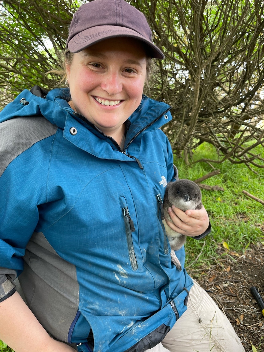 A woman outdoors, smiling while she holds a little penguin in her hand.