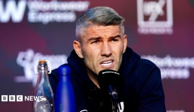 A man in his 30s, with grey hair and wearing a blue hoody, speaks into a black microphone at a press conference.