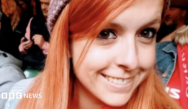 Danielle is shown smiling and wearing a beanie hat in a crowd. Her hair is ginger coloured and she is looking at the camera with her brown eyes.