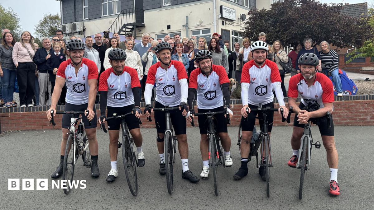 Six men in matching red and white cycling outfits sit on their bikes with the Aston Fields Social Club behind them - a two storey building with a cream base and a grey second floor. A crowd of supporters also stand behind the men.