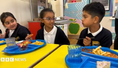 Three primary school pupils in uniform sit at bright yellow tables in a school canteen, eating lunch from blue trays with chips, fruit and dessert while talking together.