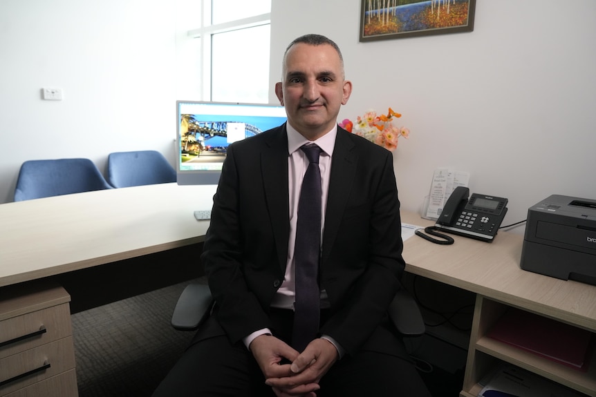 Man (Fred Betros) in a black suit sits at a desk looking at the camera.