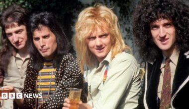 The members of Queen, holding drinks and smiling at the camera. They are wearing 1970s clothes and all have long hair.