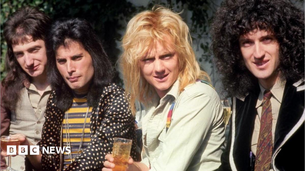 The members of Queen, holding drinks and smiling at the camera. They are wearing 1970s clothes and all have long hair.