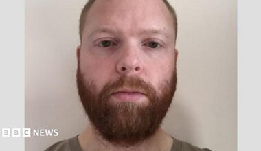 A police mugshot of Paul Chowles wearing a brown T-shirt and standing against a white background. He has a full beard and short red-brown hair, and is looking at the camera with a blank expression.