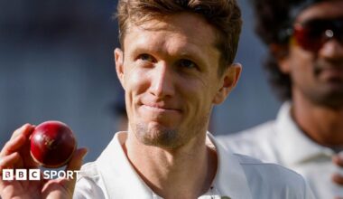 Lancashire's Will Williams holds up a red cricket ball in his right hand. He is looking up with a smile on his face