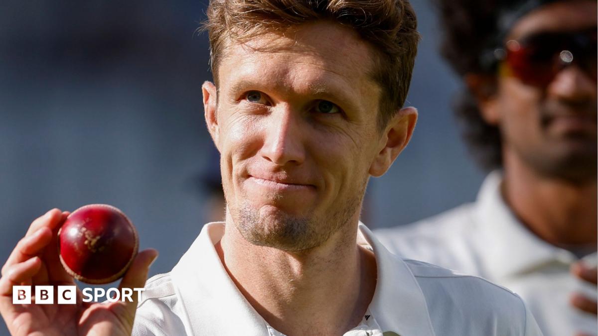 Lancashire's Will Williams holds up a red cricket ball in his right hand. He is looking up with a smile on his face