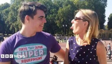 Oliver McGowan, wearing a purple t-shirt with a logo, is standing outside Buckingham Palace. He is starring at his mother Paula McGowan who is standing next to him and wearing a dark blue polka dot dress and sunglasses. She is playfully sticking her tongue out at her son and he is laughing.