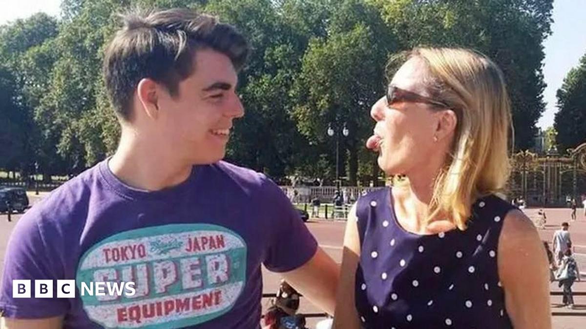 Oliver McGowan, wearing a purple t-shirt with a logo, is standing outside Buckingham Palace. He is starring at his mother Paula McGowan who is standing next to him and wearing a dark blue polka dot dress and sunglasses. She is playfully sticking her tongue out at her son and he is laughing.