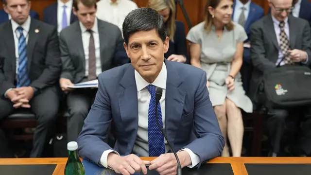 Wash sits at a desk with a mic and water bottle in front of him. There are people in the gallery behind him