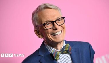 Bob Mackie wears a navy blue blazer, blue and white striped shirt and a multi-coloured tartain style bow-tie. He's smiling and there's a bright pink background.