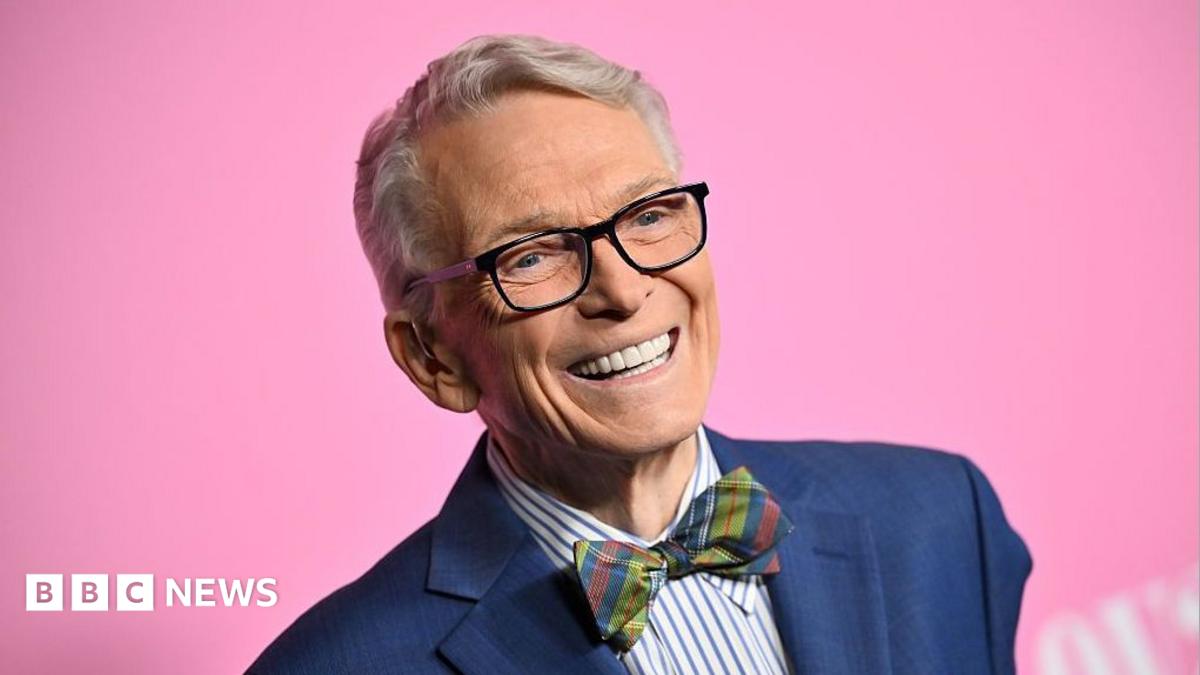 Bob Mackie wears a navy blue blazer, blue and white striped shirt and a multi-coloured tartain style bow-tie. He's smiling and there's a bright pink background.