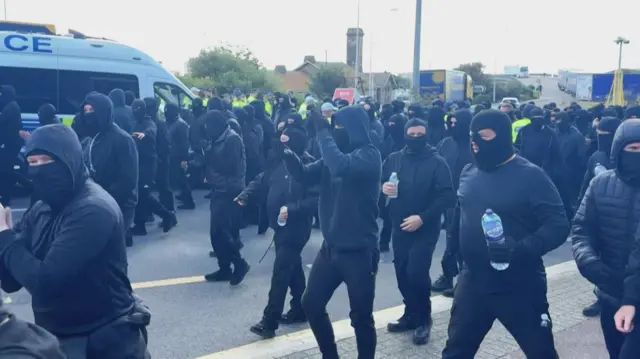 A large group of protesters walk from right to left along the road. They're all wearing black clothing and balaclavas over their heads