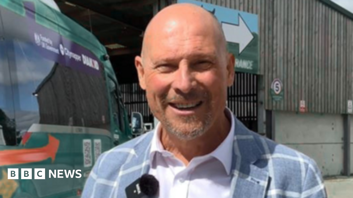 Adam Kent in a plaid blue blazer and white shirt, he is stood in front of a bus at a bus station