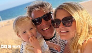 Lily is on a beach with her dad, Jonny, and her mum, Rosie.  The beach and sea are in the background.  Lily is in a sundress; Jonny and Rosie are wearing sunglasses and are smiling at the camera.