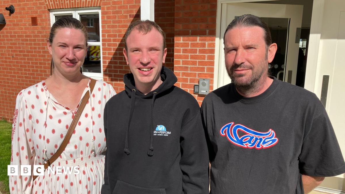 A woman in a white dress with red spots, stands next to a man in a black hoodie who stands next to a man in a black t-shirt with "vans" written in the centre in blue writing. They are standing in front of a brick building.