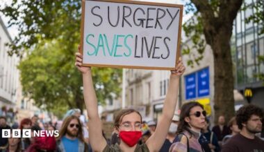 A crowd of people walking down a busy shopping street. The young person at the front of the group has tied-back brown hair and glasses and is wearing a red face mask. Their sign, which they are holding above their head, says: "Surgery saves lives".