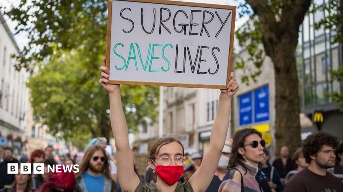 A crowd of people walking down a busy shopping street. The young person at the front of the group has tied-back brown hair and glasses and is wearing a red face mask. Their sign, which they are holding above their head, says: "Surgery saves lives".